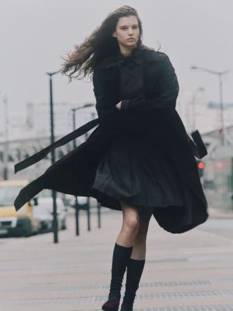 A young woman in a dark coat and knee-high socks poses on a city street in overcast weather, with a blurry vehicle in the background.