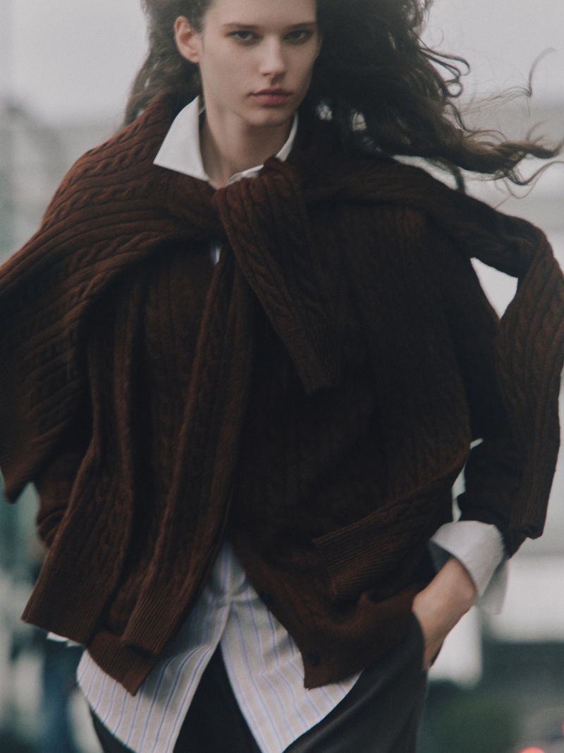A young, slender woman with wavy dark hair wears a white button-down shirt and dark pants, with a brown sweater draped across her shoulders outdoors.