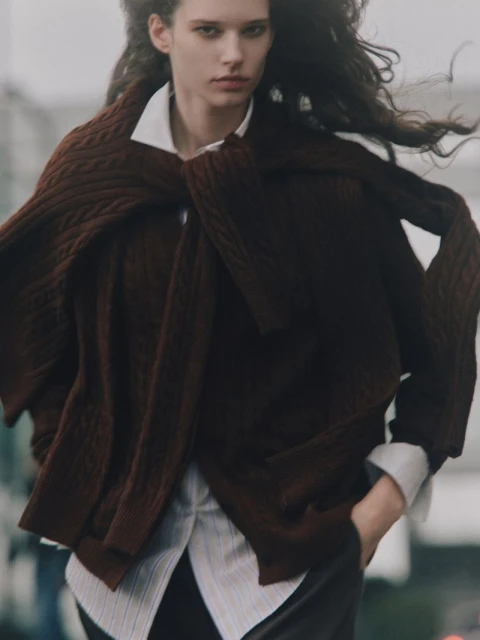 A young, slender woman with wavy dark hair wears a white button-down shirt and dark pants, with a brown sweater draped across her shoulders outdoors.