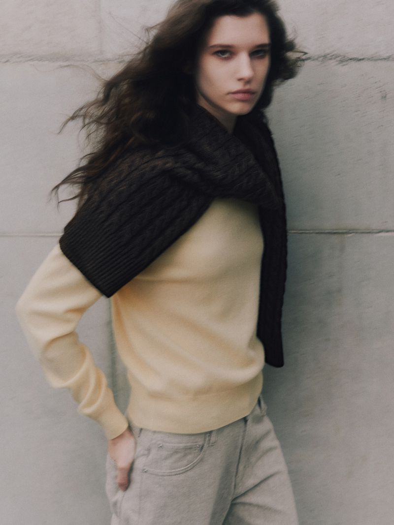 A young woman with dark hair stands in front of a concrete wall, wearing a yellow sweater, gray jeans, and a brown, knitted neck wrap.