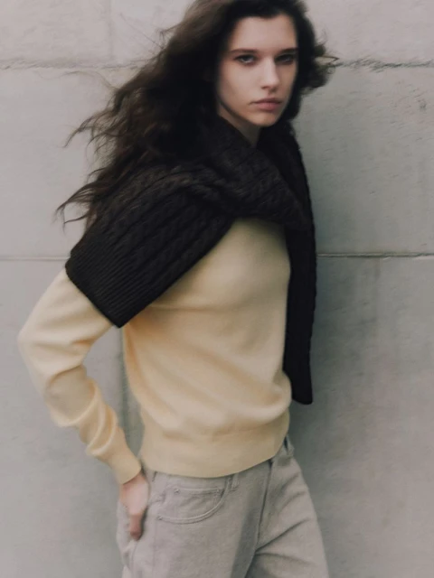 A young woman with dark hair stands in front of a concrete wall, wearing a yellow sweater, gray jeans, and a brown, knitted neck wrap.