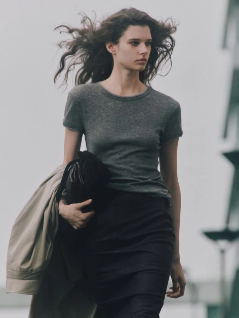 Fashion shot of a young adult woman with wavy hair in a gray t-shirt and dark skirt, holding a beige coat, standing outdoors.