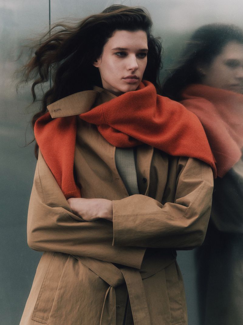 Portrait of a fashionable adult female with long brown hair wearing a tan trench coat and orange scarf outdoors in diffused lighting.
