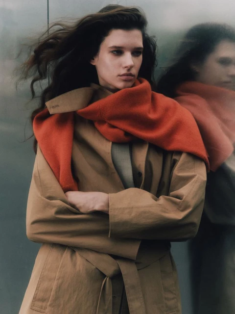 Portrait of a fashionable adult female with long brown hair wearing a tan trench coat and orange scarf outdoors in diffused lighting.