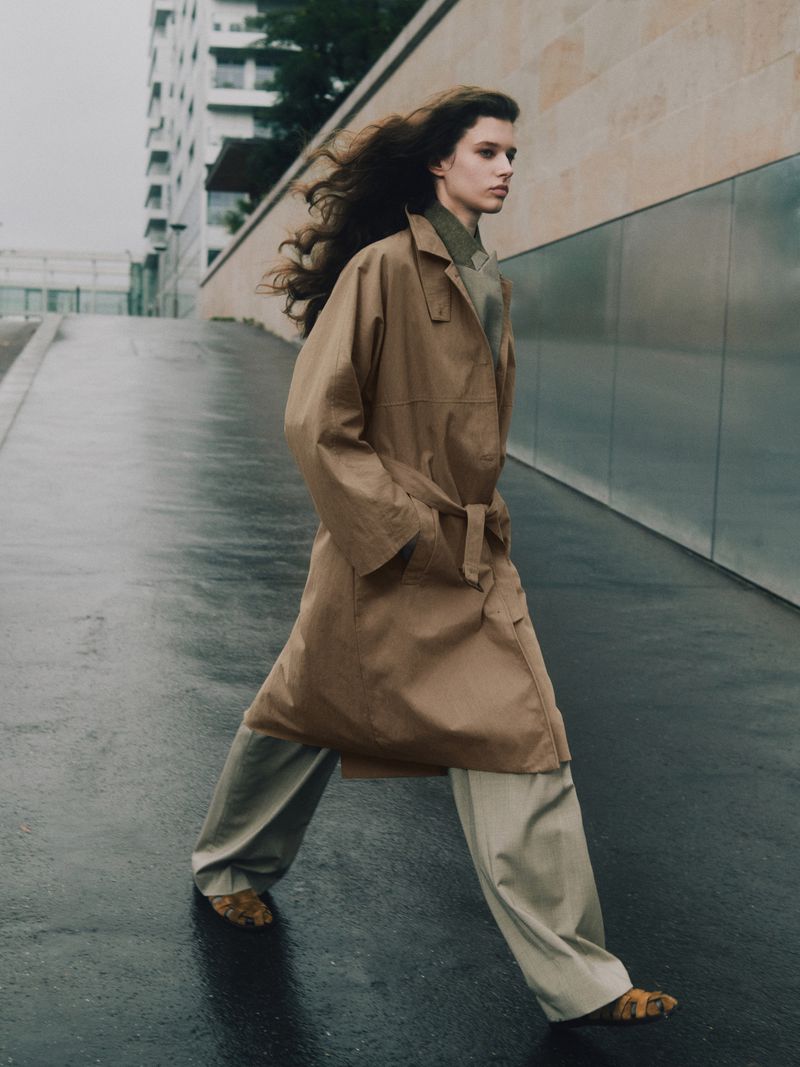 Fashionable adult female model walks on a wet street in a tan trench coat and khaki pants. Tall buildings in background.