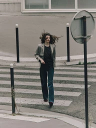 Model walking across a pedestrian crosswalk wearing a grey blazer, black top, and blue jeans
