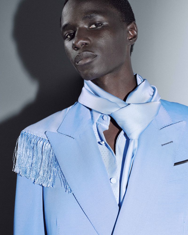 Portrait of a Black male fashion model in a light blue suit jacket with fringe and a matching scarf, set against a gradient grey background.