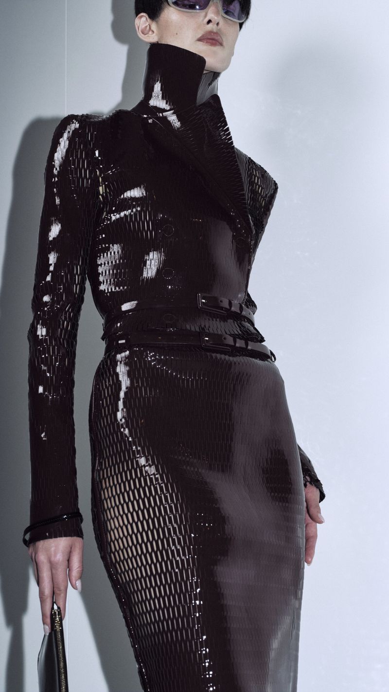 A woman models a long-sleeved, shiny brown dress with sunglasses, holding a black clutch, against a plain background in a fashion shot.