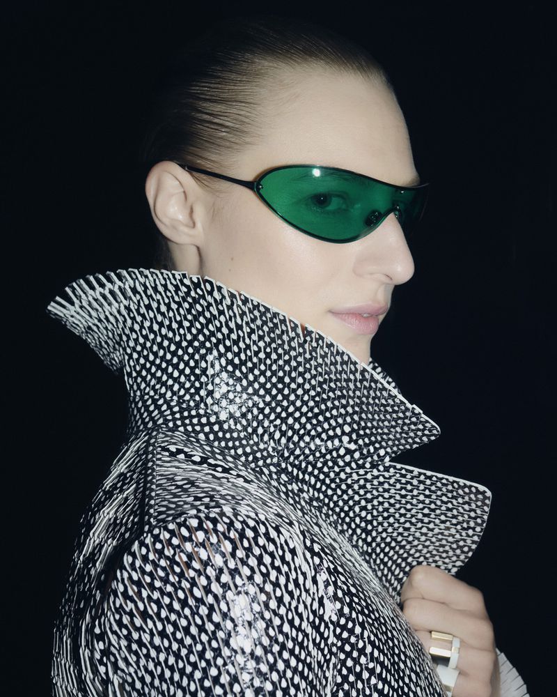 Profile shot of a person with green sunglasses and a black and white patterned jacket, against a dark background in a studio setting.