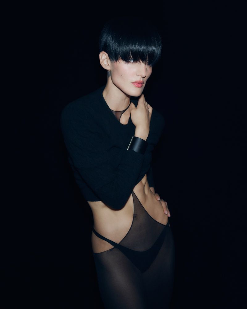 Woman with short black hair poses in semi-sheer black dress against dark backdrop. The image has a high-fashion editorial aesthetic.