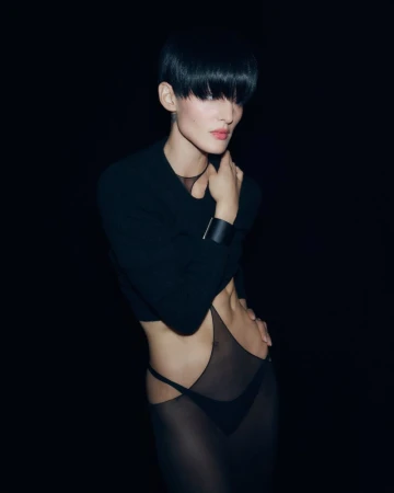Woman with short black hair poses in semi-sheer black dress against dark backdrop. The image has a high-fashion editorial aesthetic.