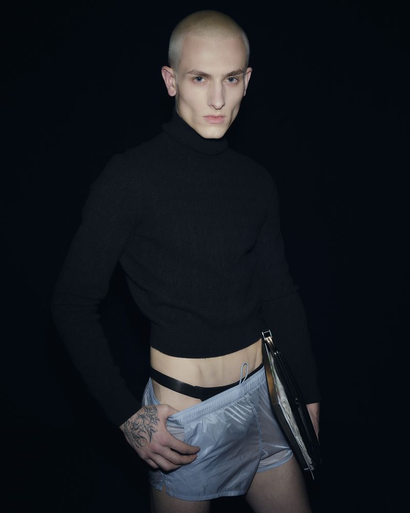 Close-up studio portrait of a fair-skinned, bald adult male wearing a black turtleneck and shiny silver shorts against a black backdrop.