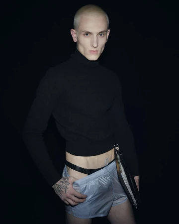 Close-up studio portrait of a fair-skinned, bald adult male wearing a black turtleneck and shiny silver shorts against a black backdrop.