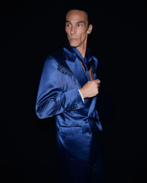 Adult male poses in a shiny royal-blue suit with fringe detail and a matching scarf against a solid black backdrop in a studio setting.