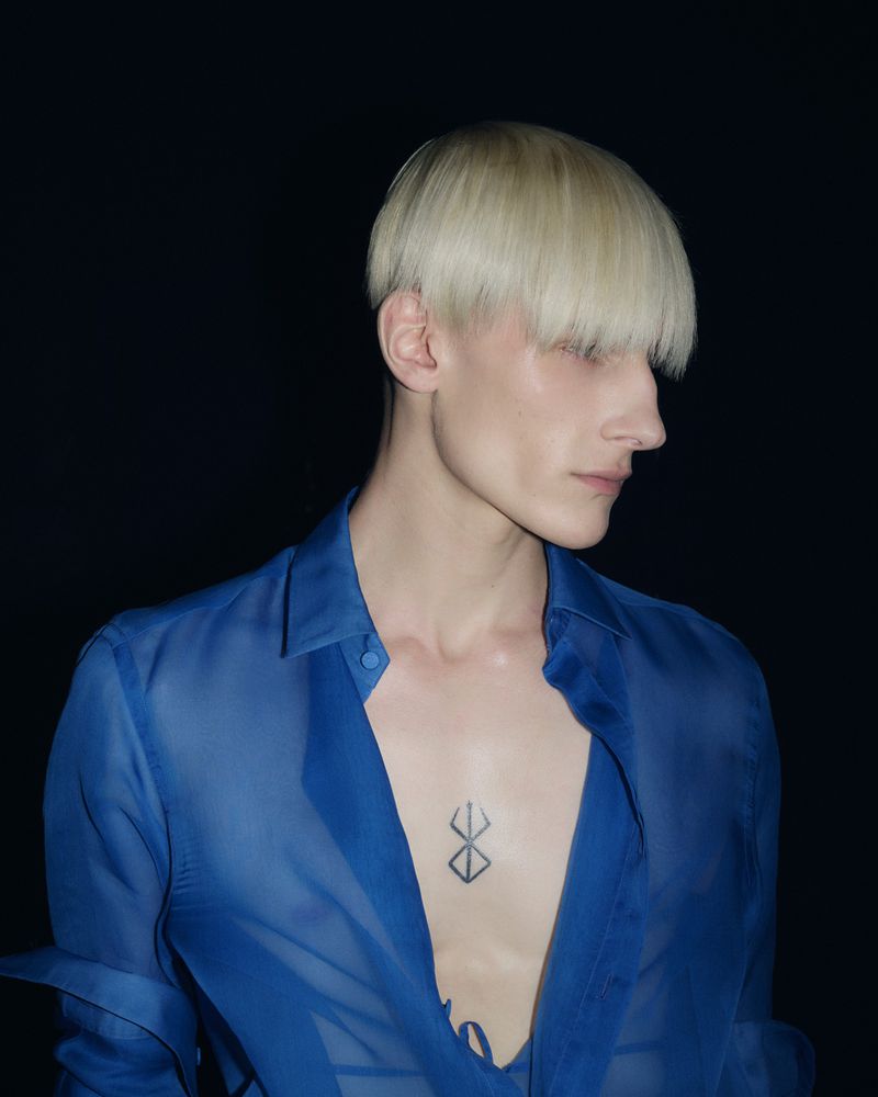 Headshot of a person with blonde hair and a sheer blue shirt against a black background, showcasing a modern fashion aesthetic.