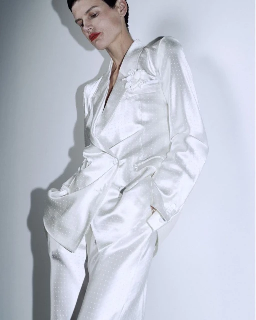 Fashion shot of an adult woman in a dotted white satin suit posing in front of a white wall, lit with studio lighting, and a medium shot.
