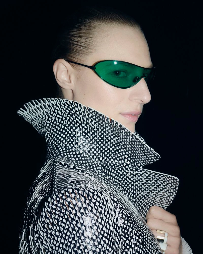 Close-up portrait of a model wearing green tinted sunglasses and a patterned black and white high collar coat against a dark background.