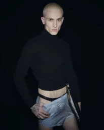 A bald male model wearing a black turtleneck and light blue shorts stands against a dark background holding a black folder.