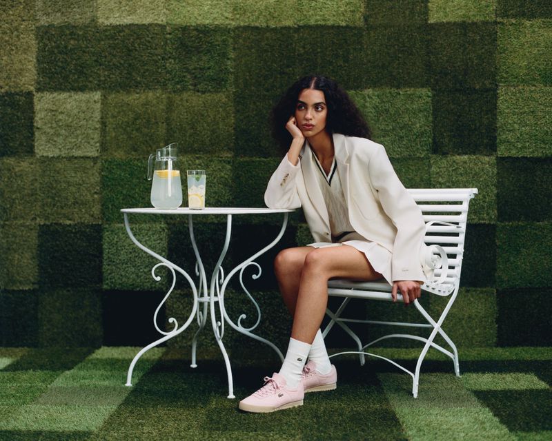 Woman poses at a white table with lemonade on a grassy checkered surface wearing a white suit and pink tennis shoes.