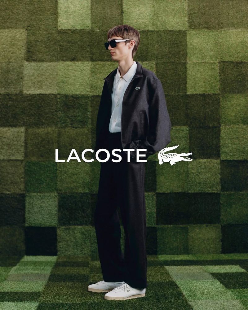 Man in Lacoste jacket and pants posing against a green geometric background, wearing sunglasses and white sneakers.