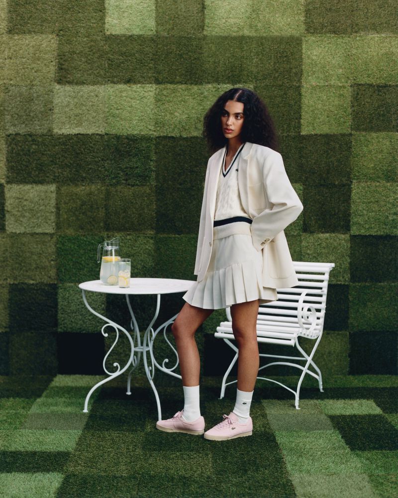A young woman wearing a cream colored tennis outfit poses in front of a green checkered background on a sunny day.