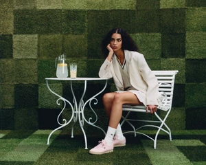 Model wearing a white blazer and pink sneakers seated at a white wire table outdoors with a tiled green backdrop.