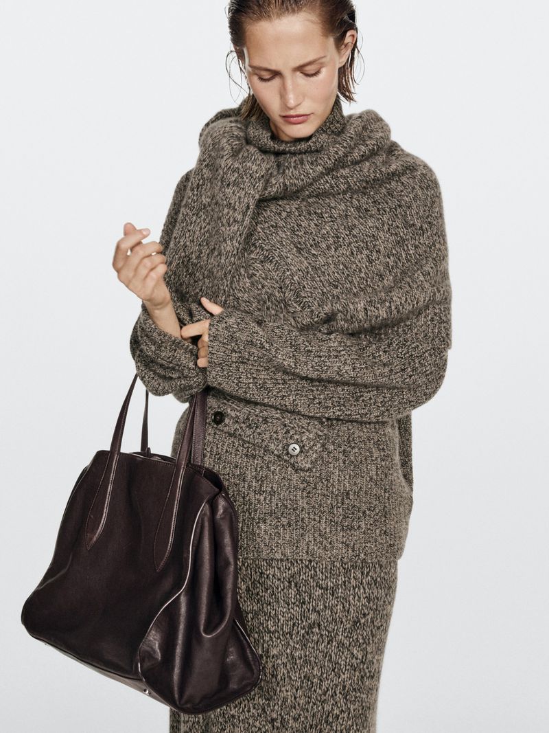 Woman wearing a wool sweater, skirt and scarf carrying a leather tote bag in a fashion shot against a white background.