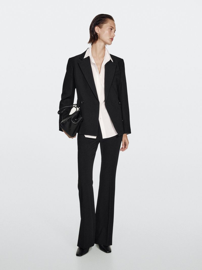 A full-length studio shot features an adult female model in a black suit and flared pants holding a black purse against a white background.