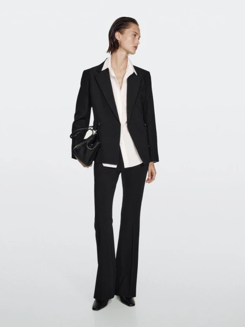 A full-length studio shot features an adult female model in a black suit and flared pants holding a black purse against a white background.