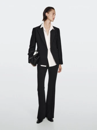 A full-length studio shot features an adult female model in a black suit and flared pants holding a black purse against a white background.