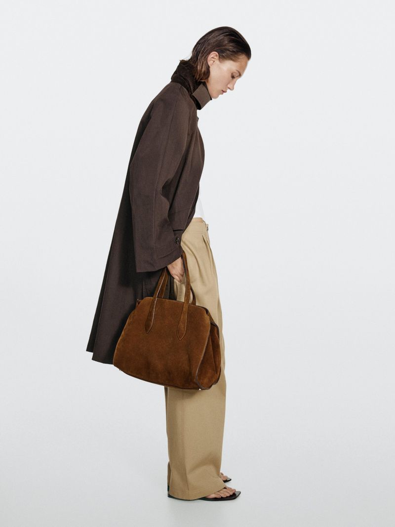 A woman wearing a brown trench coat and beige trousers, carrying a brown suede handbag, in a neutral, bright studio setting.