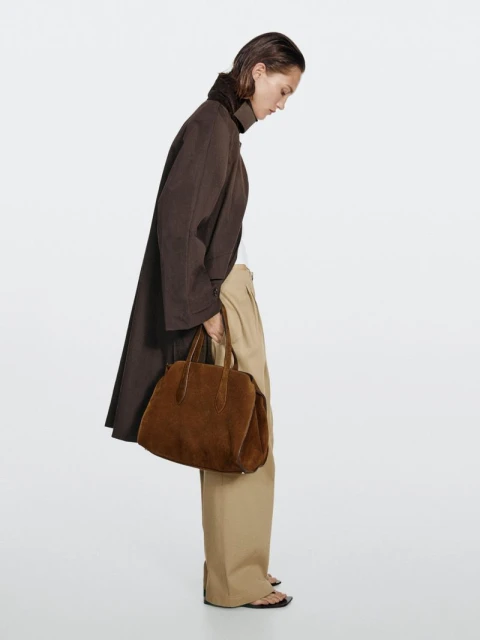 A woman wearing a brown trench coat and beige trousers, carrying a brown suede handbag, in a neutral, bright studio setting.