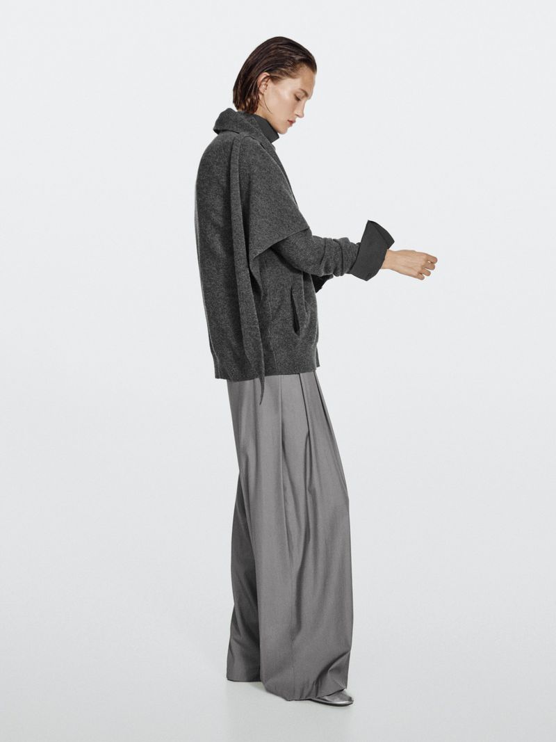 Indoor fashion shot of an adult female model in gray, posing in a sweater and wide-leg pants against a clean white backdrop.