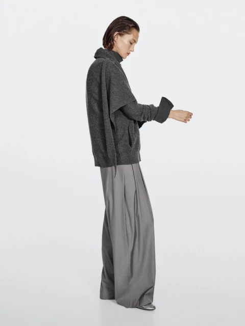 Indoor fashion shot of an adult female model in gray, posing in a sweater and wide-leg pants against a clean white backdrop.