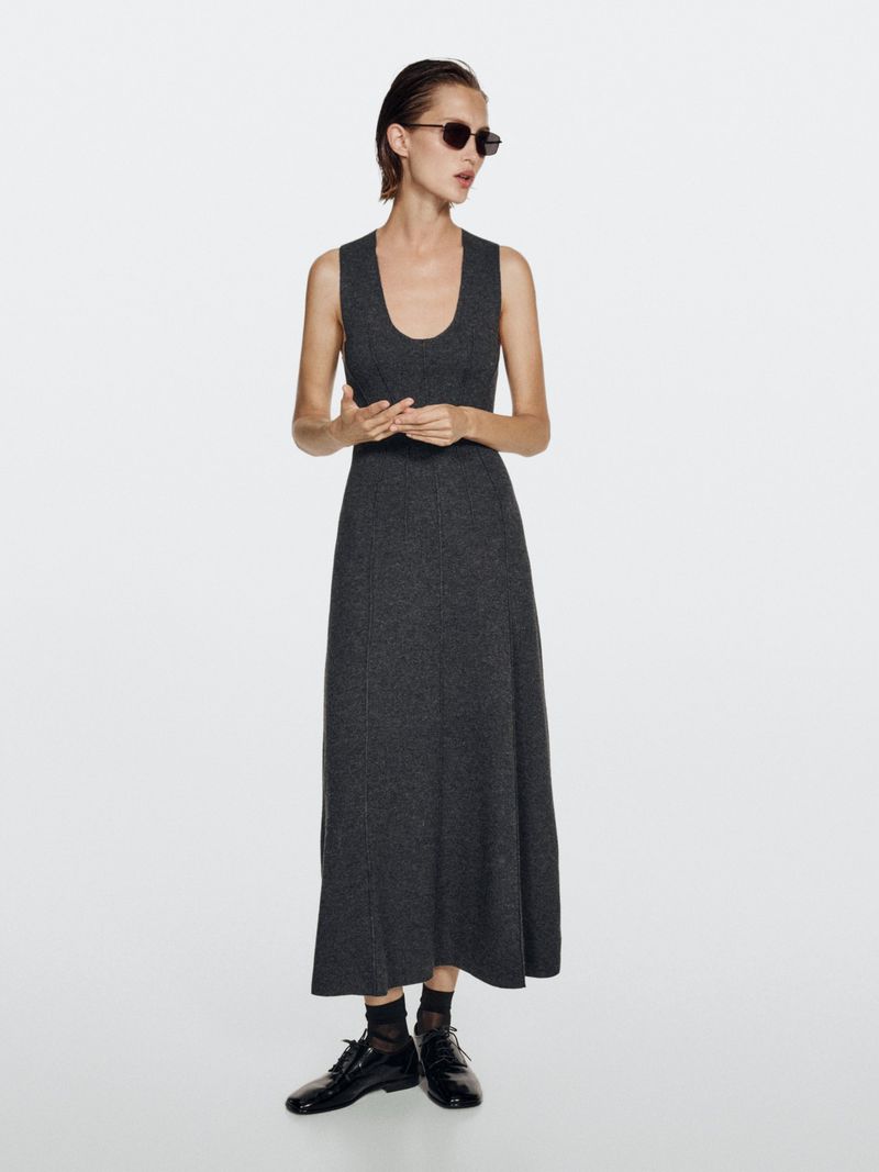Adult woman poses wearing a gray dress, sunglasses, and black shoes in a studio with a bright white backdrop and even lighting.