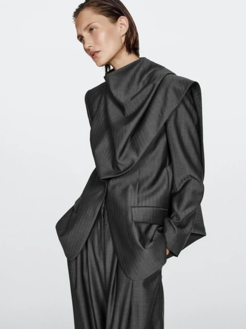Woman in a structured, draped grey suit posing against a white backdrop in a studio setting. Fashion editorial, portrait shot.