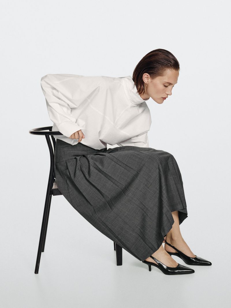 Model Vika Evseeva seated on a black chair wearing a white blouse and grey pleated skirt for Massimo Dutti Back to Office project.