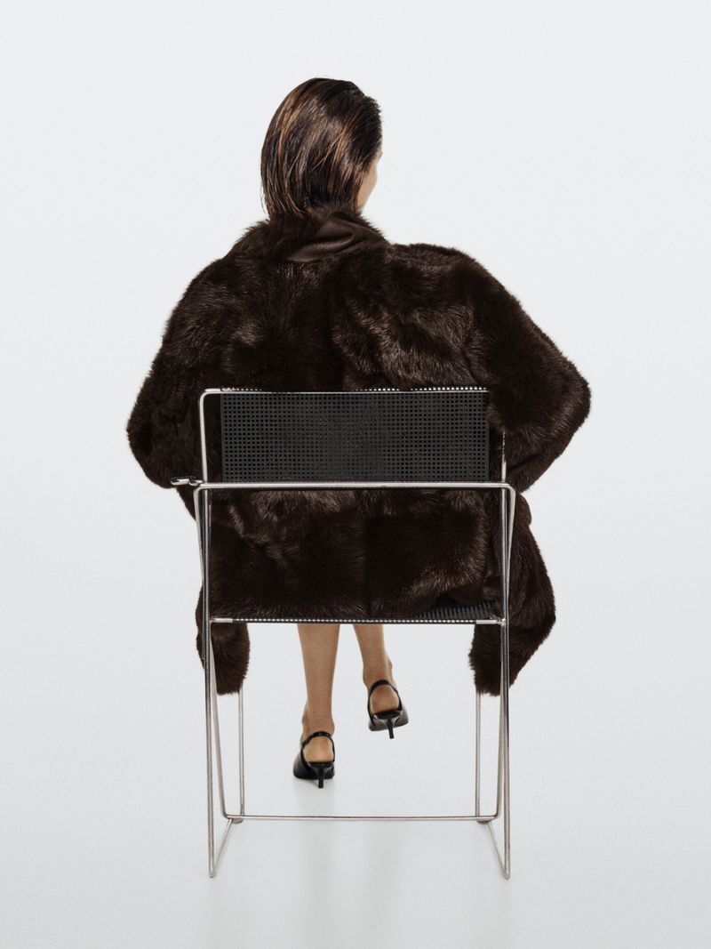 Model Vika Evseeva seen from behind wearing a dark brown coat while seated backward on a metal frame chair against a white background.