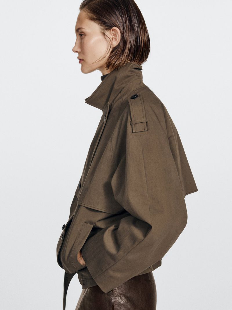 Side profile of a woman wearing an olive-green jacket and brown skirt against a white backdrop in a studio fashion setting.