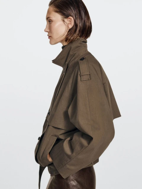 Side profile of a woman wearing an olive-green jacket and brown skirt against a white backdrop in a studio fashion setting.