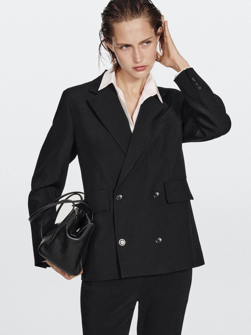 Model wearing a black double-breasted blazer and white shirt holding a black leather handbag against a plain background.