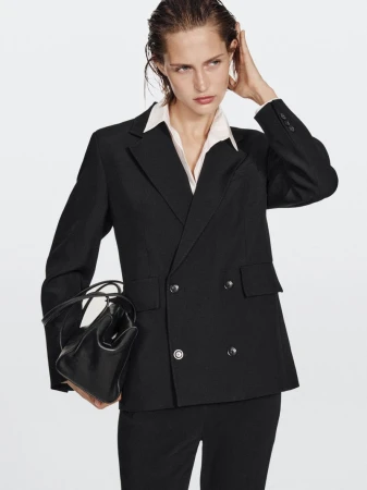 Model wearing a black double-breasted blazer and white shirt holding a black leather handbag against a plain background.