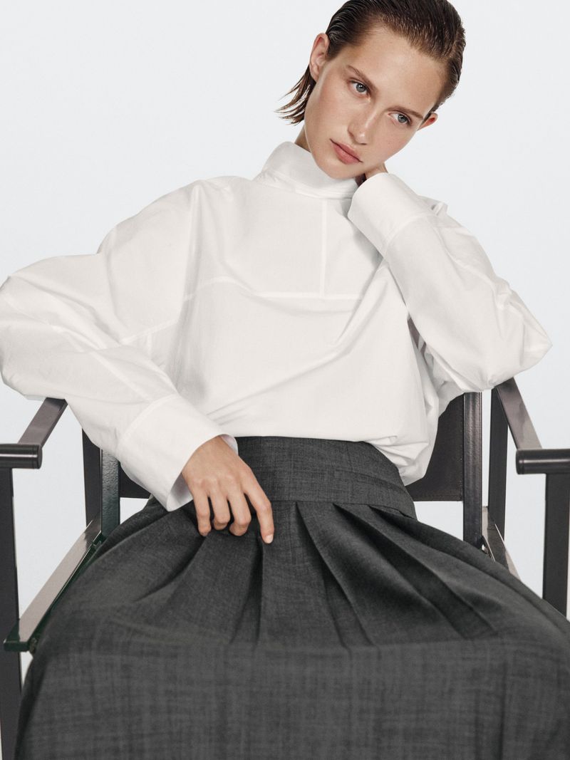 Fashion model in a white long-sleeved top and pleated skirt poses on a black chair against a white backdrop in a studio setting.
