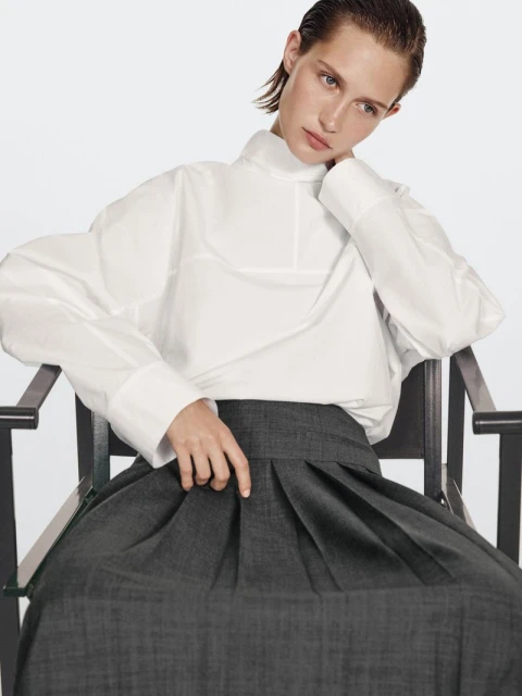 Fashion model in a white long-sleeved top and pleated skirt poses on a black chair against a white backdrop in a studio setting.