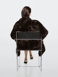 Model Vika Evseeva seen from behind wearing a dark brown coat while seated backward on a metal frame chair against a white background.