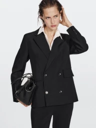 Model wearing a black double-breasted blazer and white shirt holding a black leather handbag against a plain background.