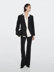 Model wearing a black blazer, white shirt, and black trousers while holding a black handbag against a plain background