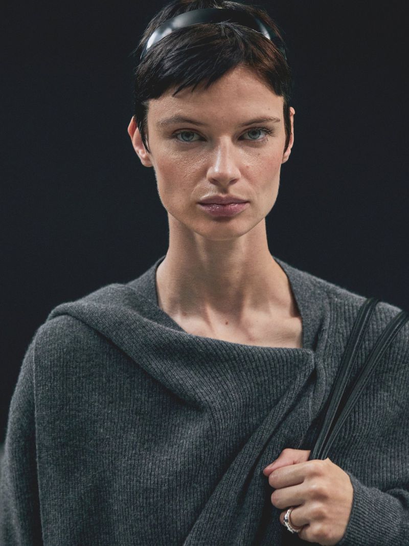Portrait of an adult female with short dark hair and a black headband, wearing a grey wrap sweater indoors against a dark backdrop.