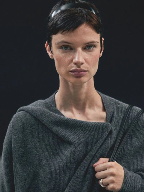 Portrait of an adult female with short dark hair and a black headband, wearing a grey wrap sweater indoors against a dark backdrop.