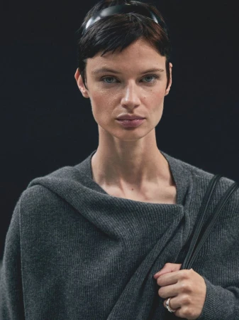 Portrait of an adult female with short dark hair and a black headband, wearing a grey wrap sweater indoors against a dark backdrop.